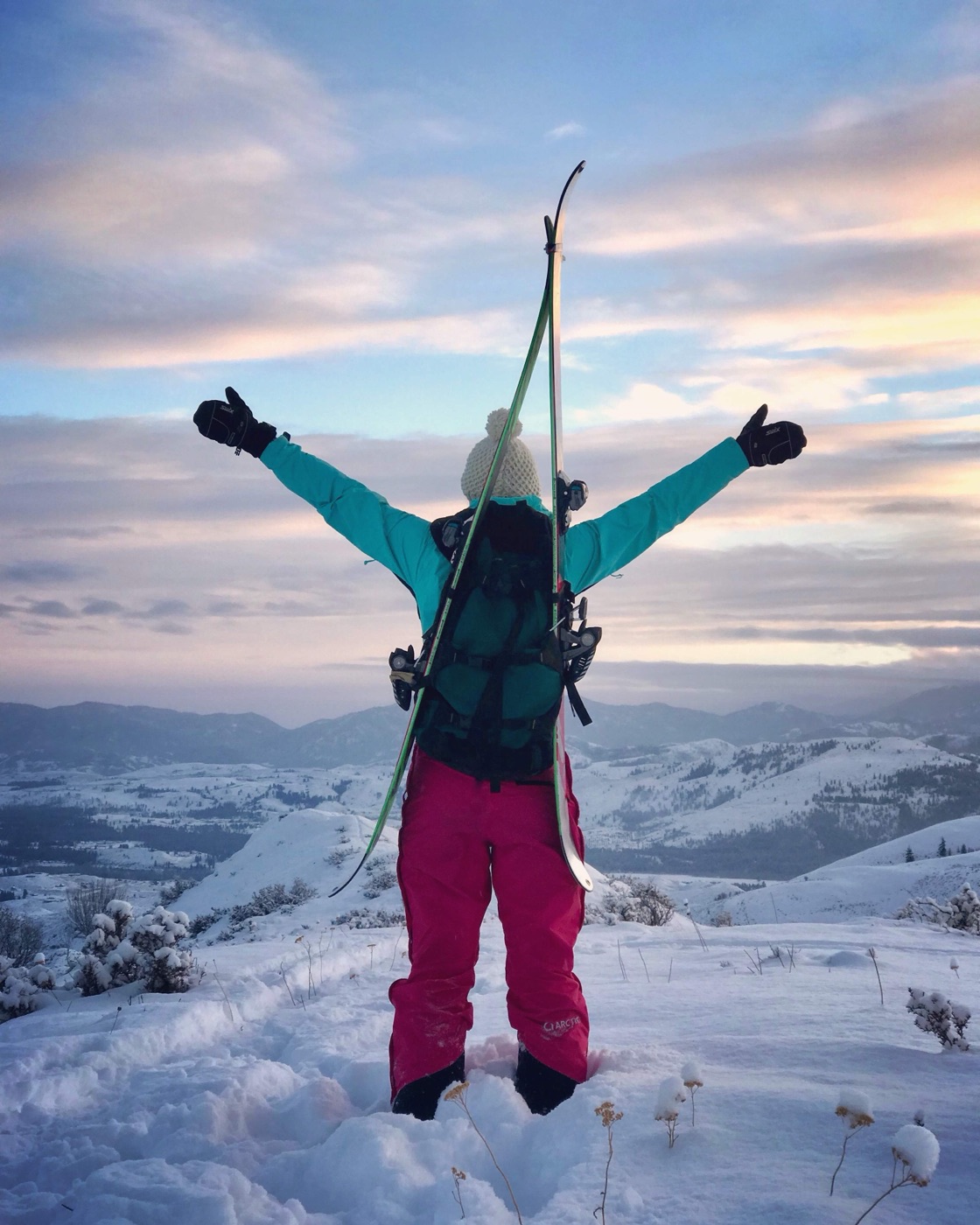 A backcountry skier on a snowy summit at sunset, skis on her pack, arms raised to a pink and blue sky over the mountains